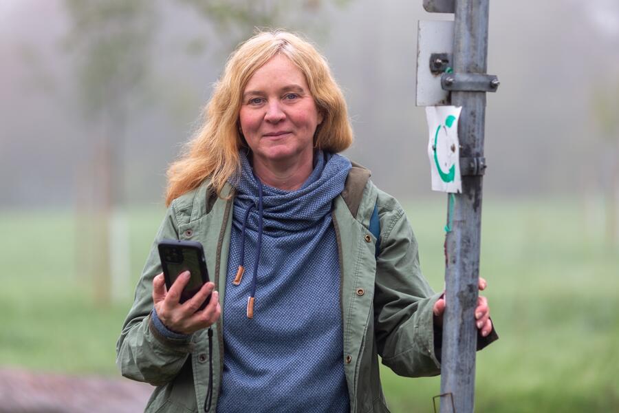 Eine Frau steht an einem Schild mit dem Symbol der Hessischen Apfelwein- und Obstwiesenroute. In der Hand hlt sie ien Smartphone.